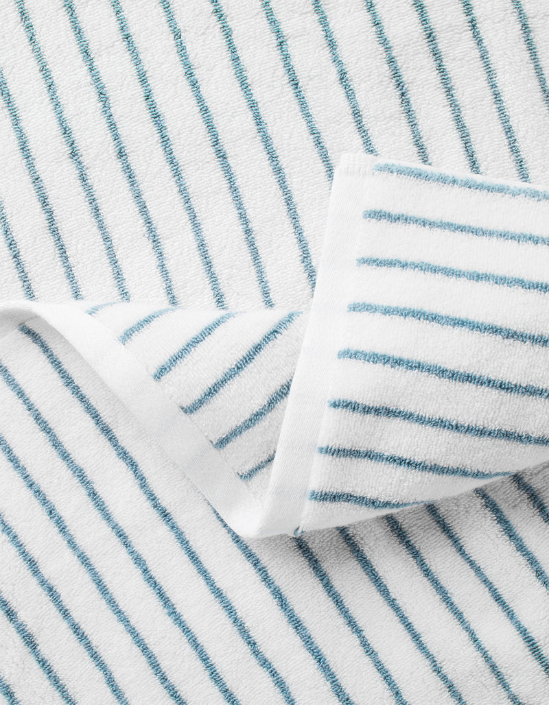 A close-up of the Cozy Earth Complete Striped Luxe Bath Bundle shows a soft, textured white towel with thin blue stripes and a folded corner that reveals the striped pattern on both sides.
