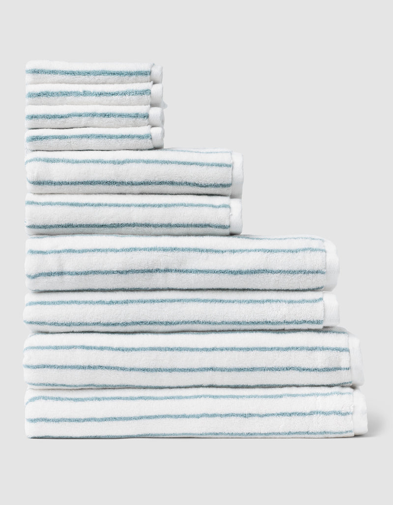 The Complete Striped Luxe Bath Bundle by Cozy Earth features neatly stacked white towels with thin blue stripes arranged in a stepped formation against a plain light gray background.