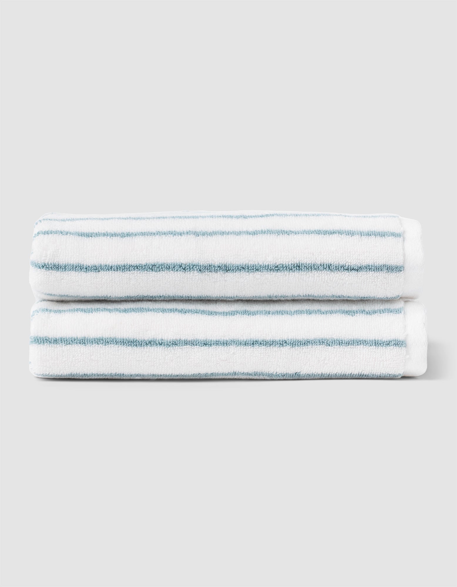 Two Striped Luxe Hand Towels by Cozy Earth, featuring thin teal horizontal stripes on white, are neatly folded and stacked against a light gray background. 