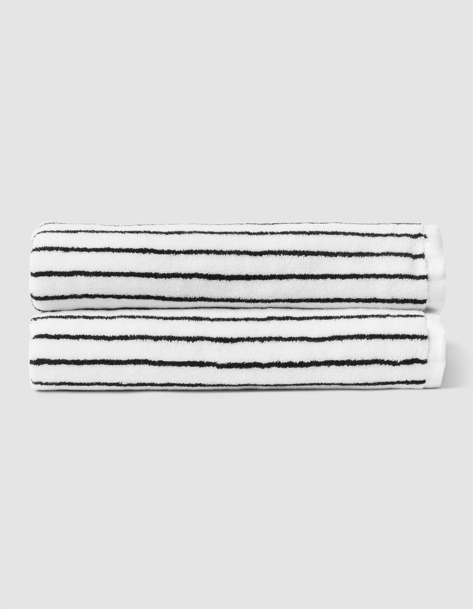 Two Striped Luxe Bath Sheets by Cozy Earth, featuring thin black horizontal stripes on white, are neatly rolled and stacked against a plain light gray background. 
