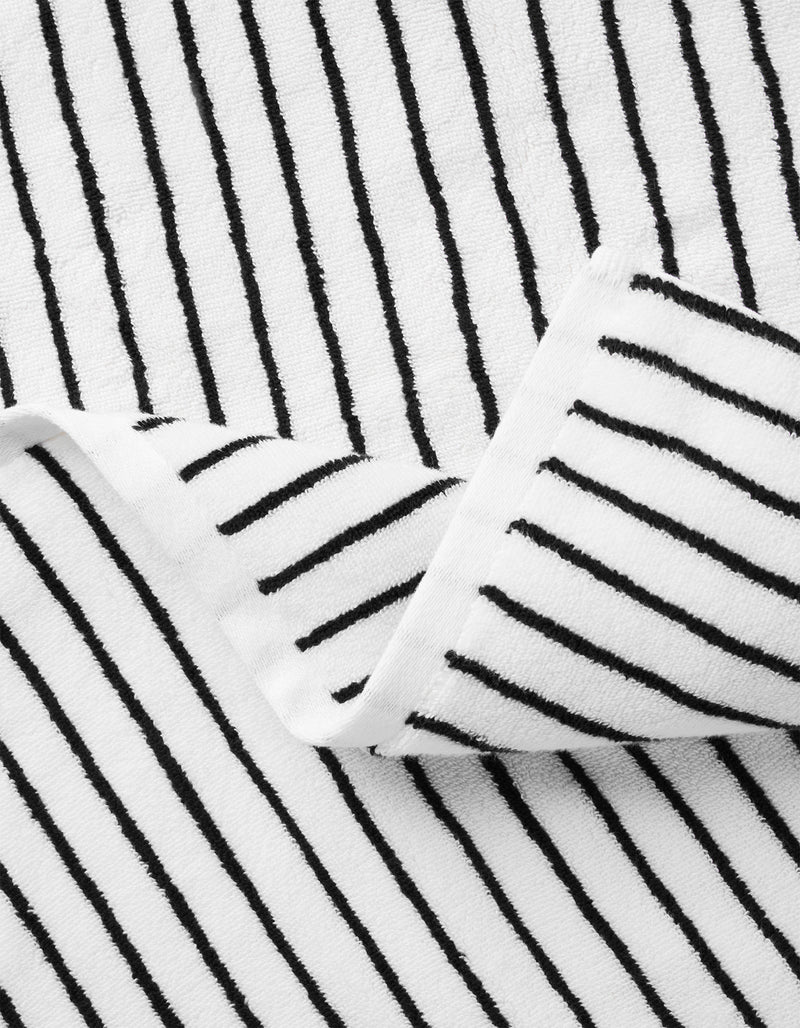 A close-up of Cozy Earth’s Complete Striped Luxe Bath Bundle shows white fabric with thin black vertical stripes; a folded corner reveals the matching striped pattern on both sides.