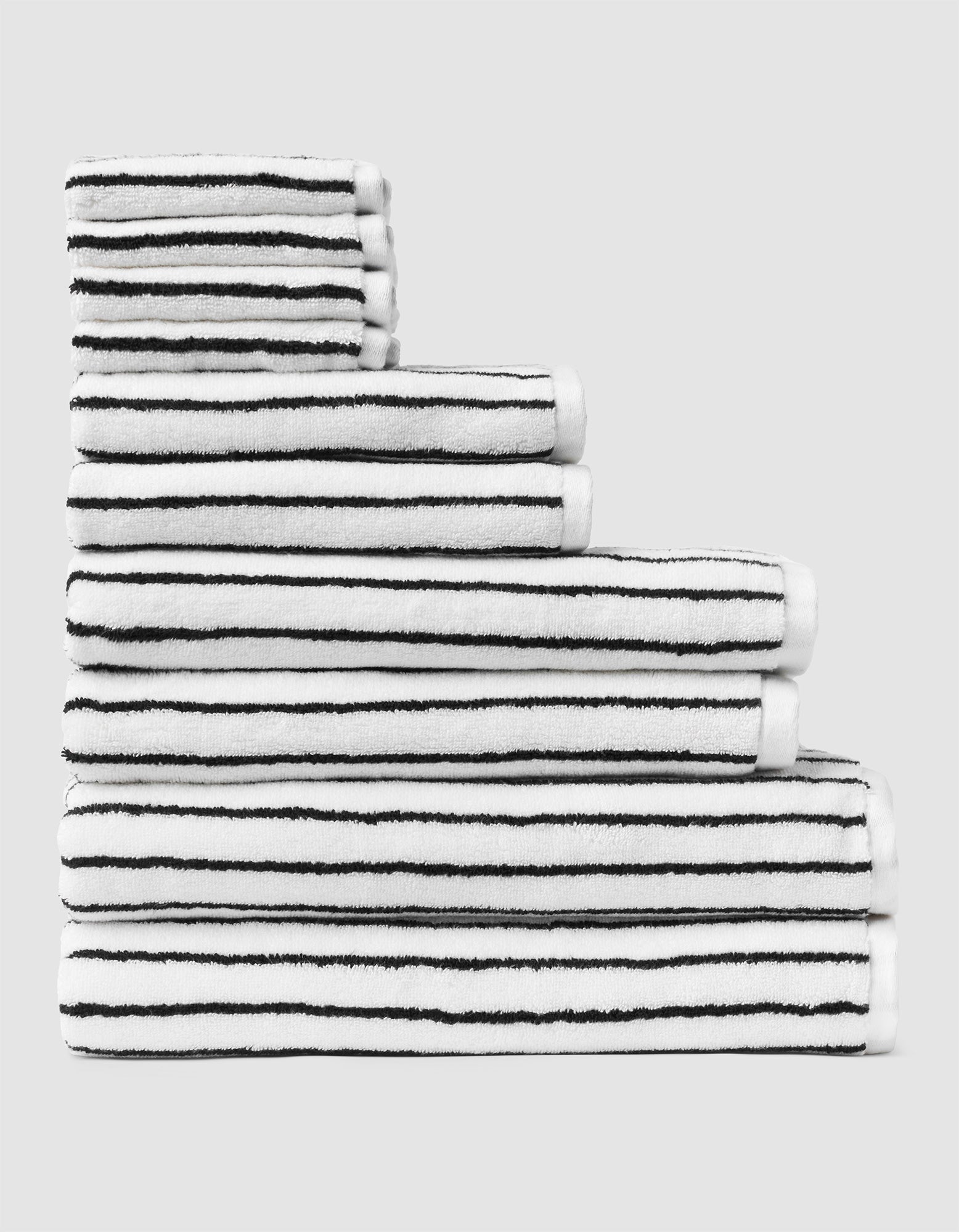 The Complete Striped Luxe Bath Bundle by Cozy Earth features neatly stacked white towels with thin black stripes, arranged in descending size order on a light gray background. 