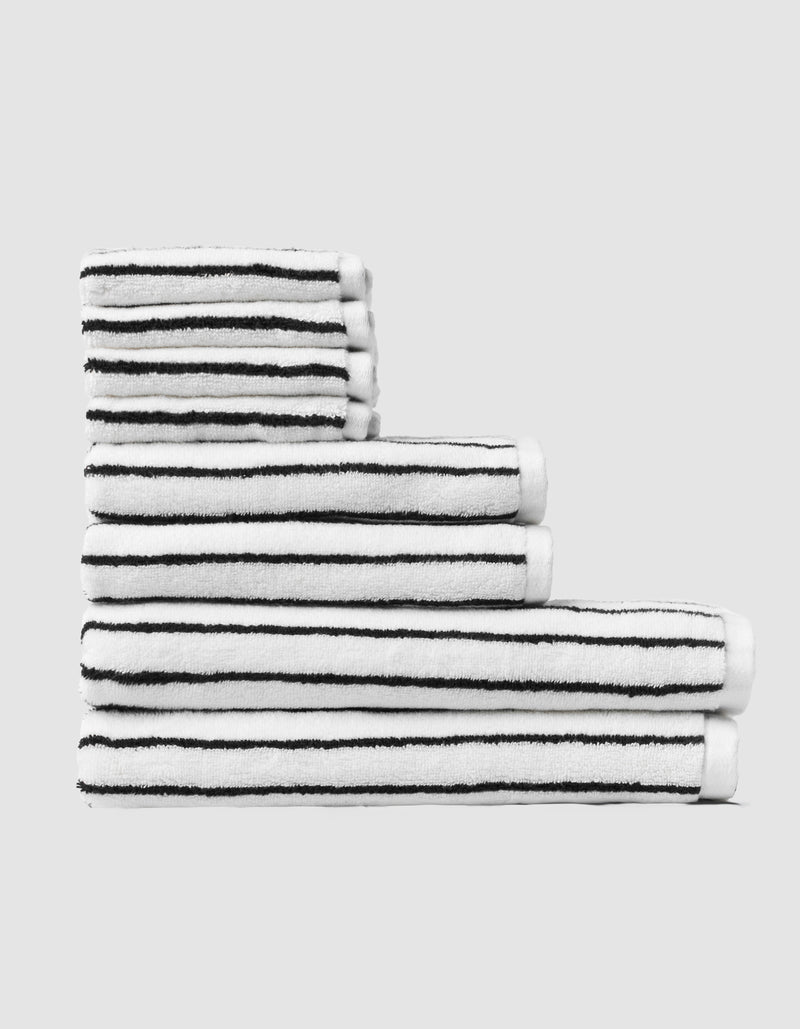 The Cozy Earth Striped Luxe Bath Towel Set features five white towels with thin black horizontal stripes, stacked in ascending order against a plain light gray background.