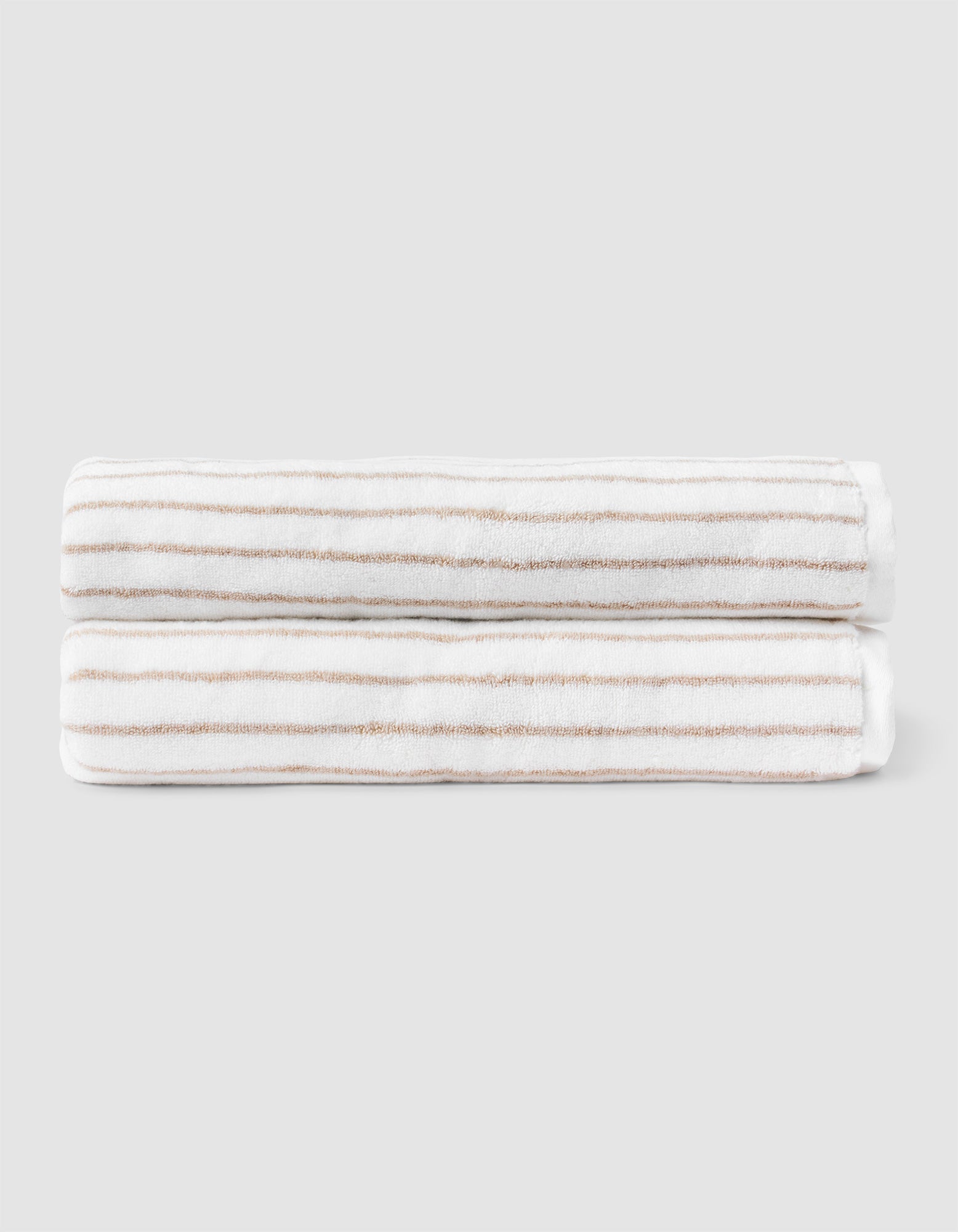 Two Striped Luxe Bath Sheets by Cozy Earth, featuring white fabric with thin, evenly spaced tan stripes, are neatly folded and stacked against a plain light gray background.  