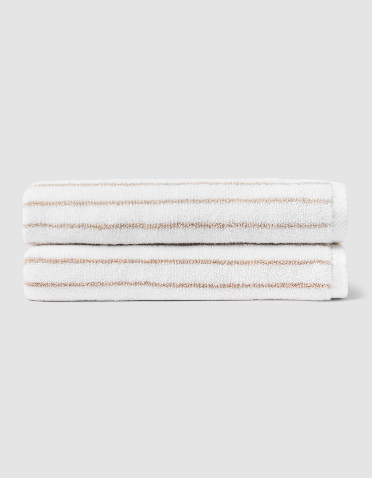 Two Striped Luxe Bath Towels by Cozy Earth, featuring thin light brown stripes on white, are stacked neatly against a plain light gray background. 