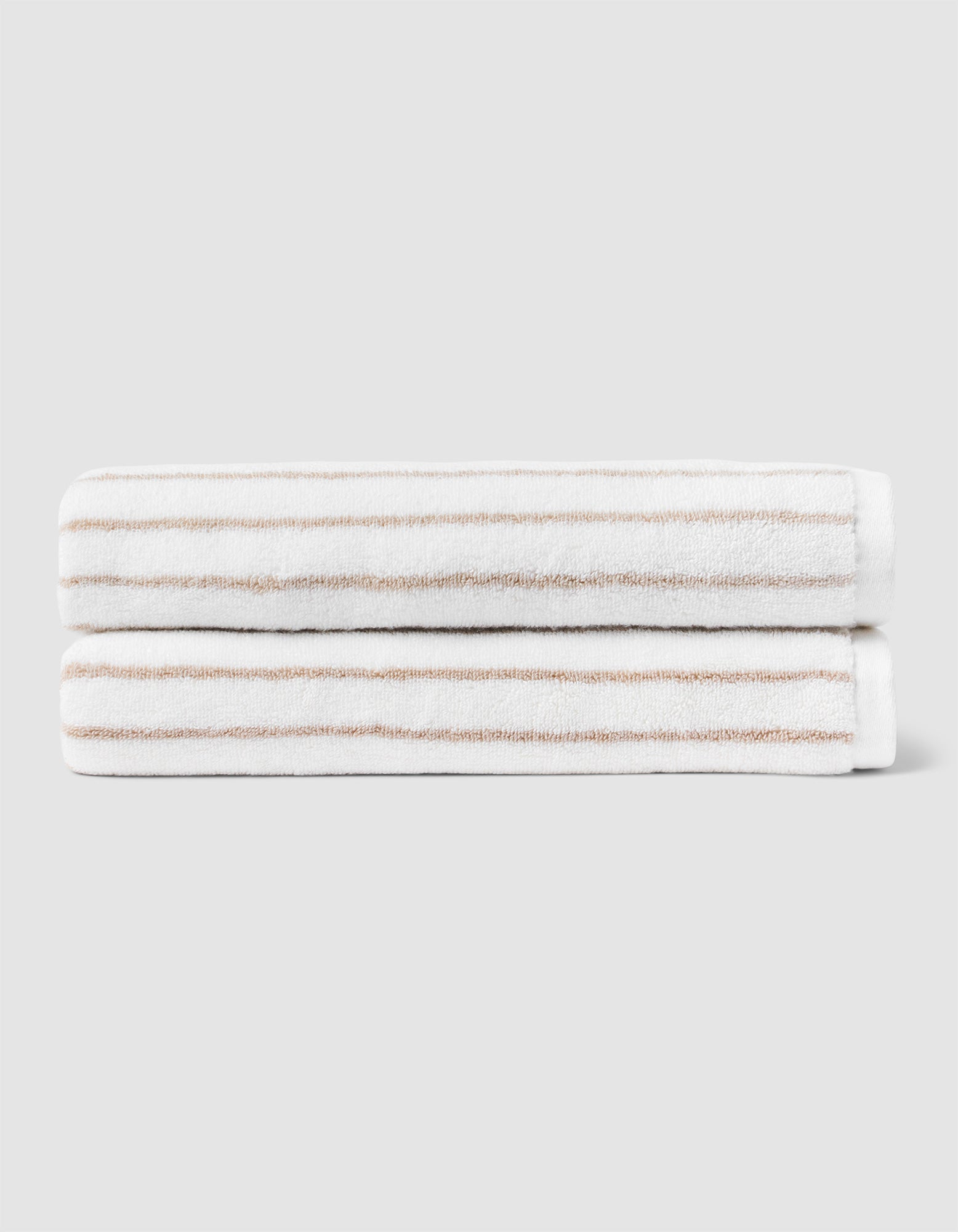 Two Striped Luxe Bath Towels by Cozy Earth, featuring thin light brown stripes on white, are stacked neatly against a plain light gray background. 
