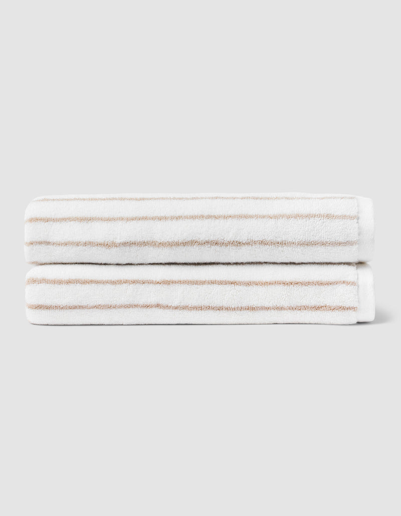 Two Striped Luxe Bath Towels by Cozy Earth, featuring thin light brown stripes on white, are stacked neatly against a plain light gray background.