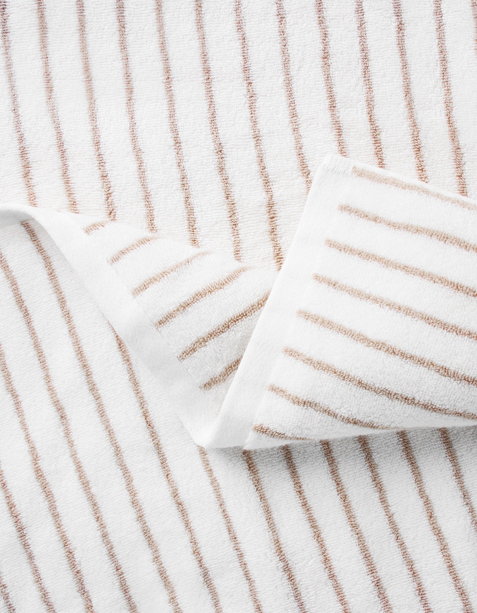 A close-up of the Cozy Earth Striped Luxe Bath Towel, featuring thin, evenly spaced brown vertical stripes on white fabric and a partial fold at one corner to display the pattern on both sides. 