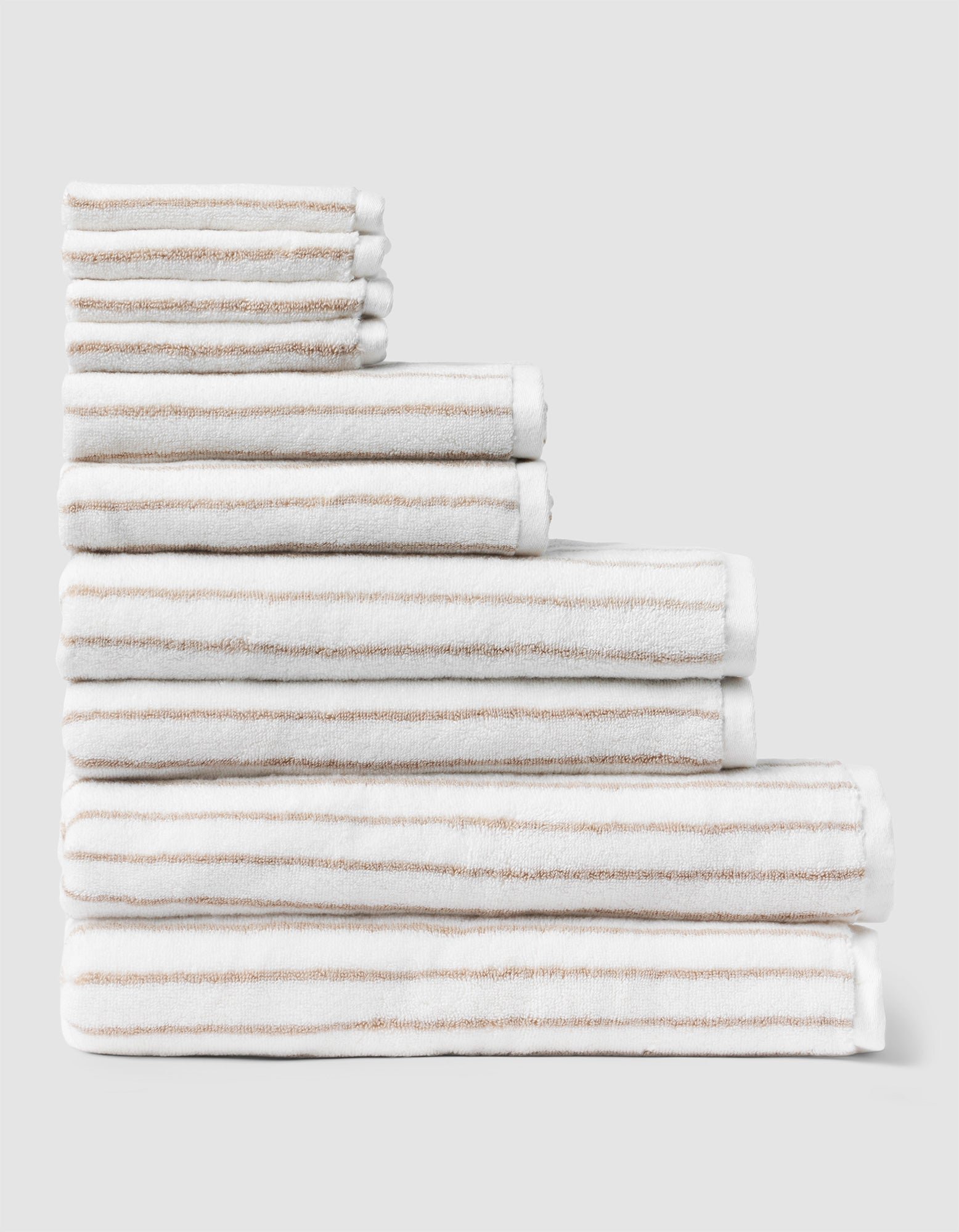 The Complete Striped Luxe Bath Bundle by Cozy Earth features neatly stacked white towels with thin beige stripes, arranged in descending size order against a plain light gray background. |Color:Sand Stripe