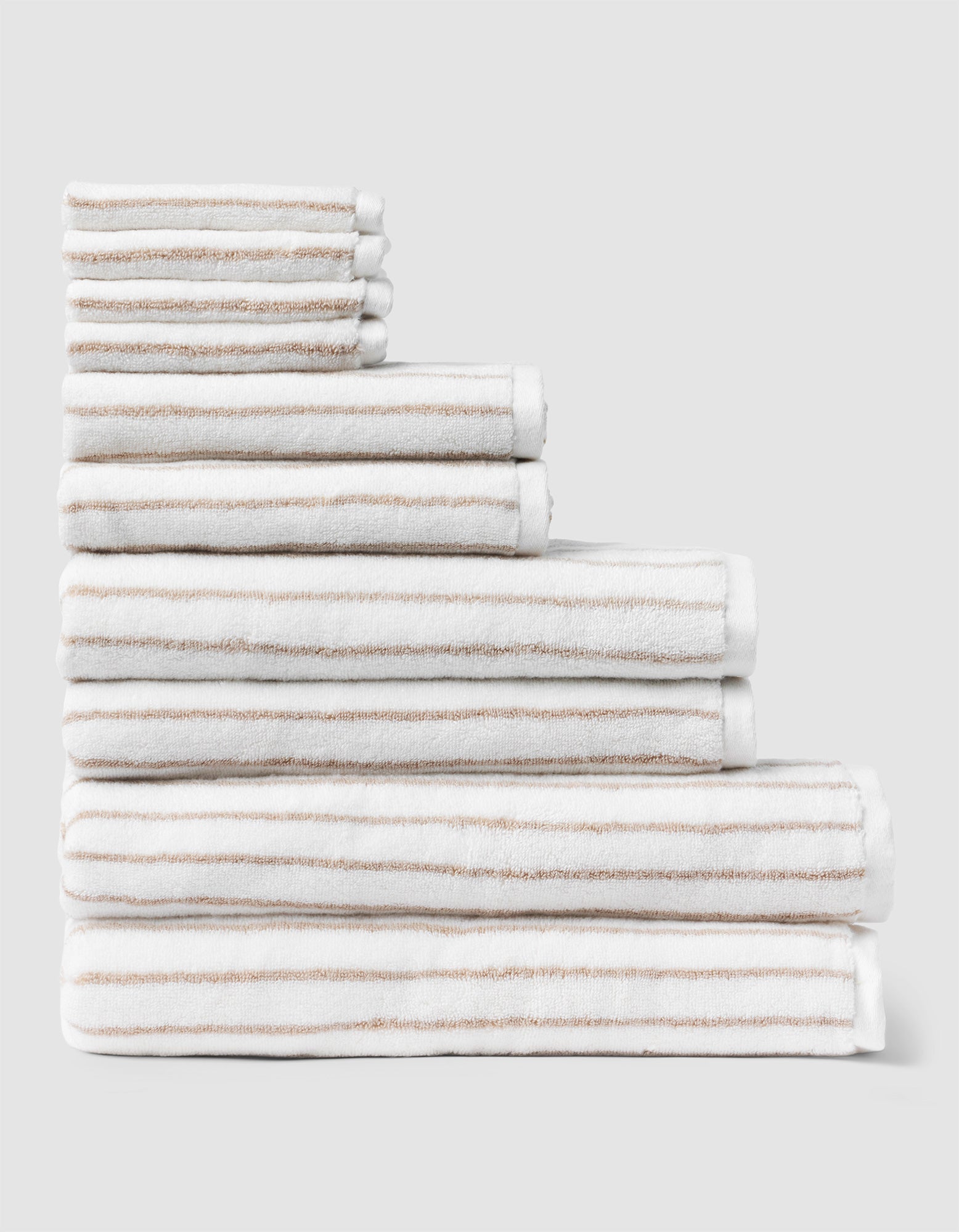 The Complete Striped Luxe Bath Bundle by Cozy Earth features neatly stacked white towels with thin beige stripes, arranged in descending size order against a plain light gray background. 