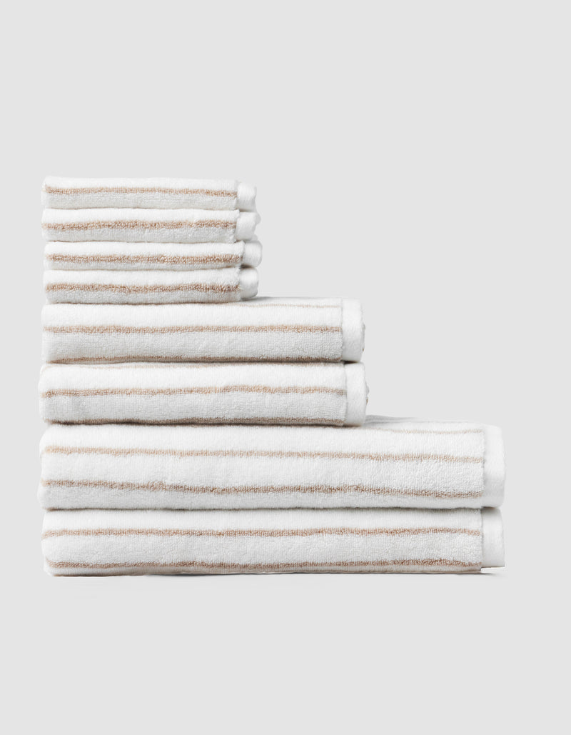 The Cozy Earth Striped Luxe Bath Towel Set features neatly folded white towels with beige stripes in three sizes, displayed against a plain light gray background.