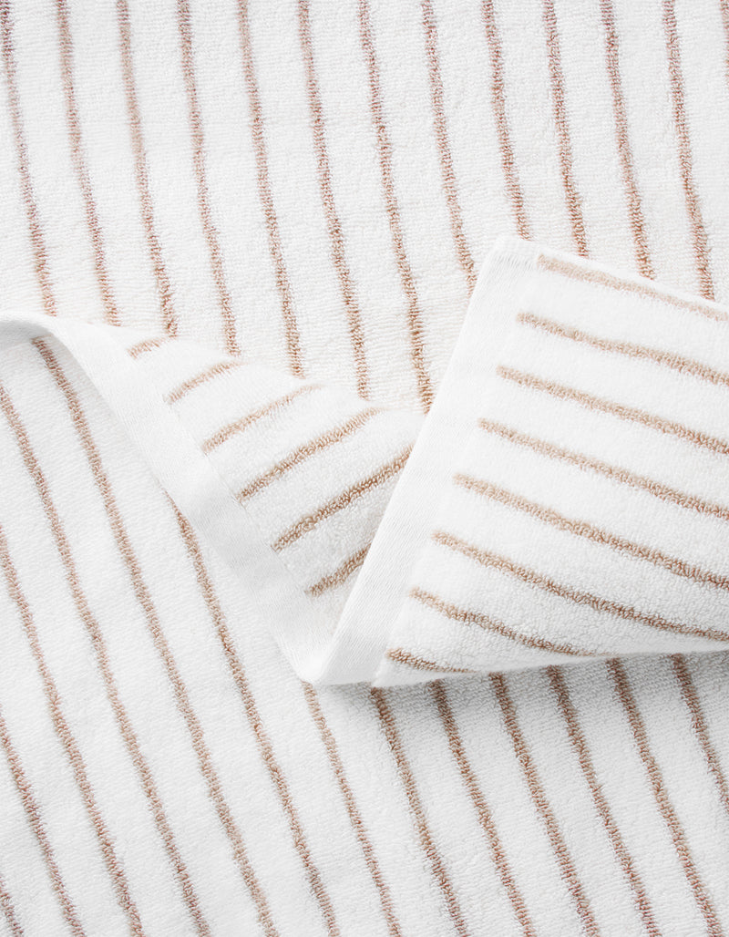 A close-up of Cozy Earth's Striped Luxe Washcloths shows evenly spaced brown stripes on white fabric, with one corner folded to display the signature striped pattern on both sides.
