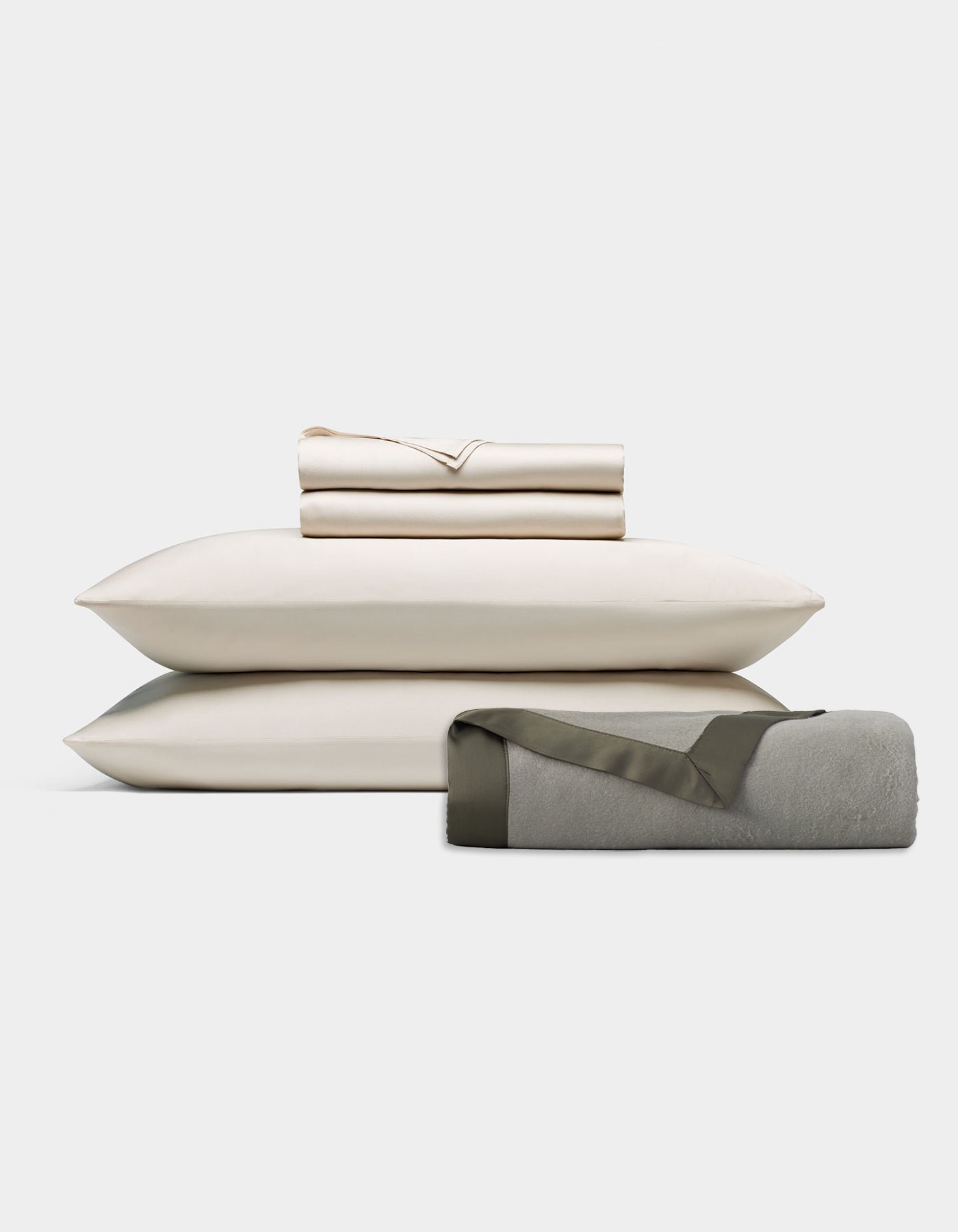 The End of Summer Bedding Bundle by Cozy Earth features cream-colored sheets and pillowcases neatly stacked on two pillows, with a rolled light gray blanket edged in dark trim, displayed against a plain white background.