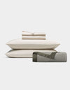 The End of Summer Bedding Bundle by Cozy Earth features cream-colored sheets and pillowcases neatly stacked on two pillows, with a rolled light gray blanket edged in dark trim, displayed against a plain white background.