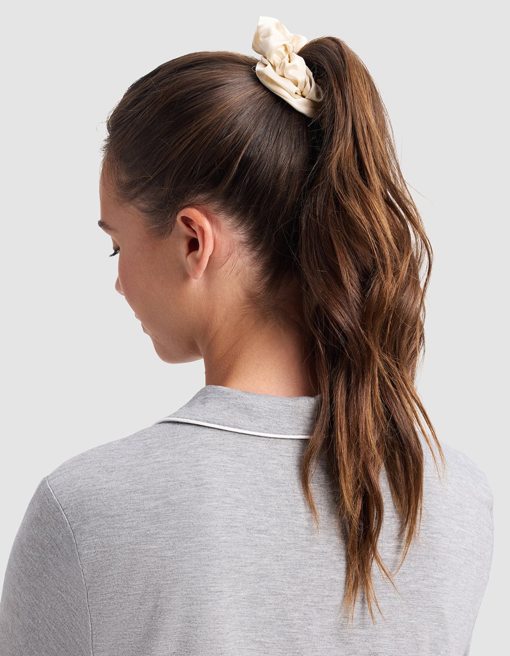 A woman with long brown hair in a ponytail wears a Cozy Earth Silk Scrunchie (Last Chance) and a light gray collared shirt, seen from behind against a plain light background.