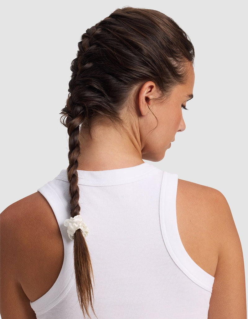 A woman with brown hair in a single braid, secured by a white Silk Skinny Scrunchie from Cozy Earth, is shown from behind wearing a white sleeveless ribbed top, facing slightly to the right.