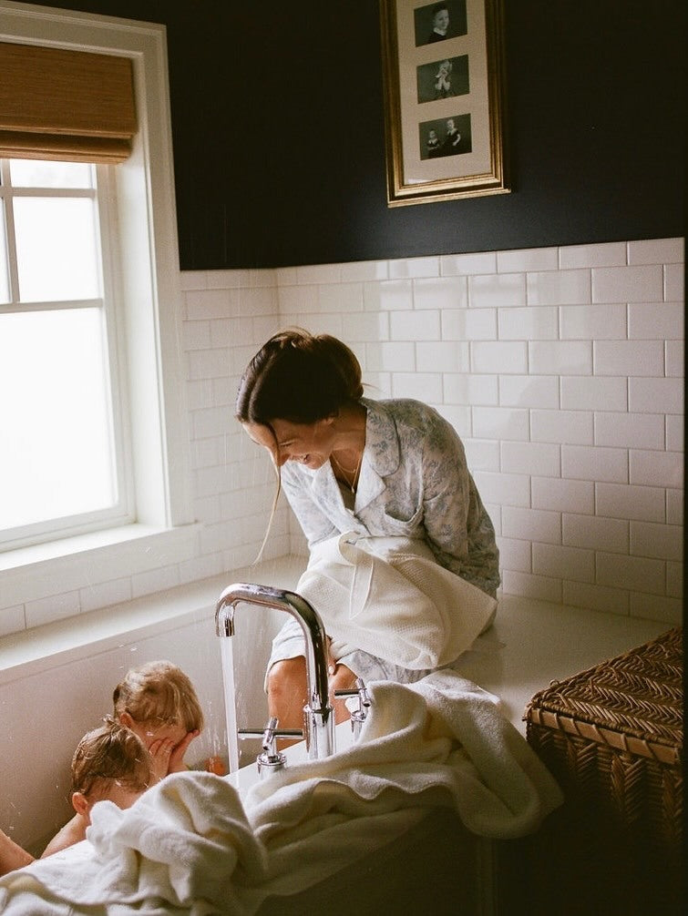 Woman and children in a bathroom with white tiled walls and a window.