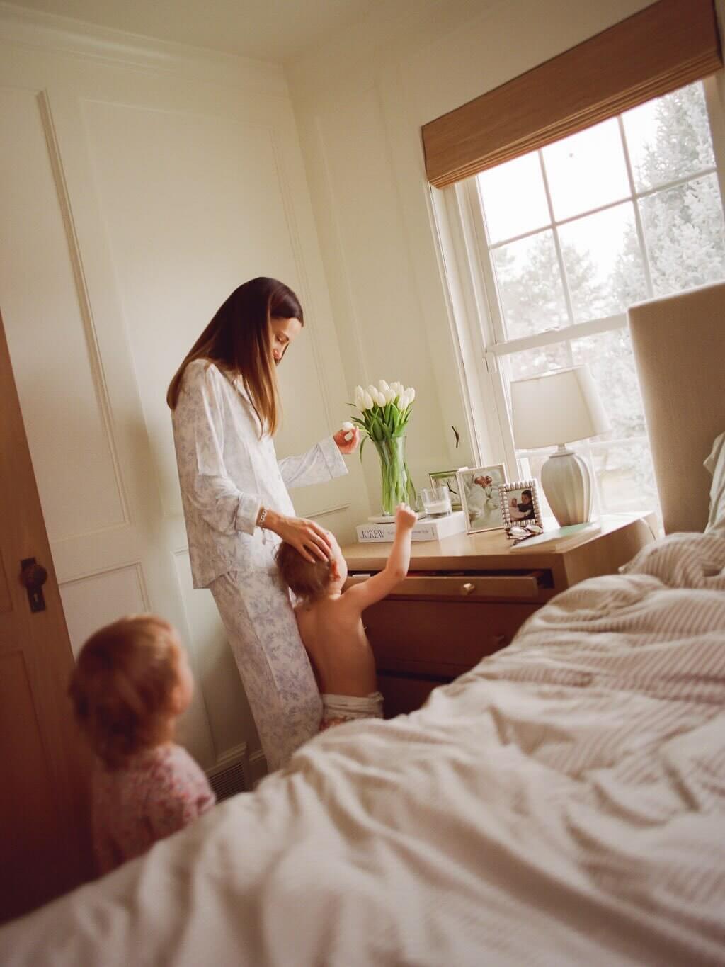 Woman in a white robe standing by a bed with a child, in a room with a large window.