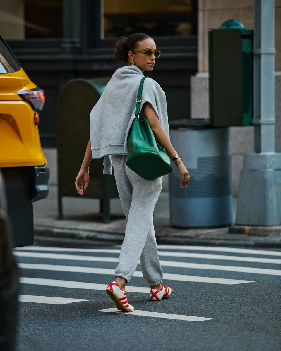 A woman walking across a city crosswalk wearing a light gray Cozy Earth Cityscape Crewneck around her shoulders and Cozy Earth Cityscape sweatpants, a green shoulder bag, and red-and-white sneakers. A yellow taxi is partially visible on the left, with urban buildings and street elements in the background.
