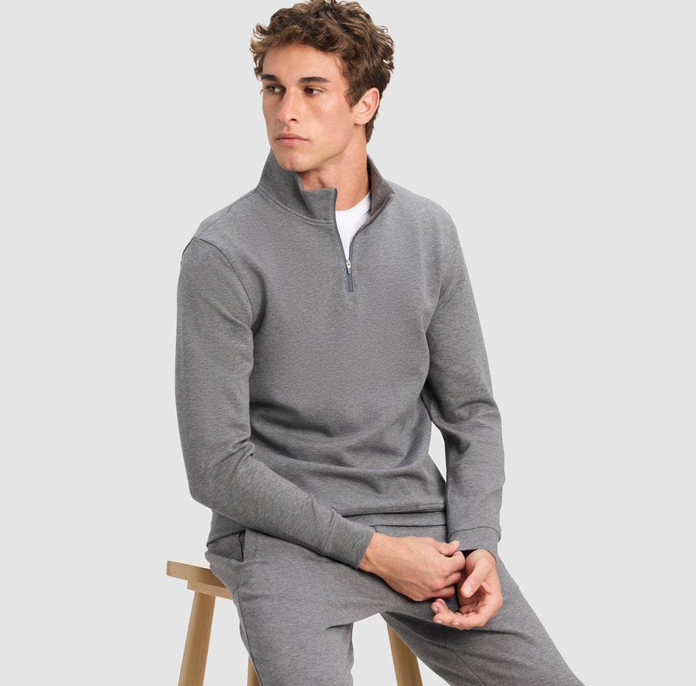 Man wearing a gray sweater and pants sitting on a wooden stool against a light gray background