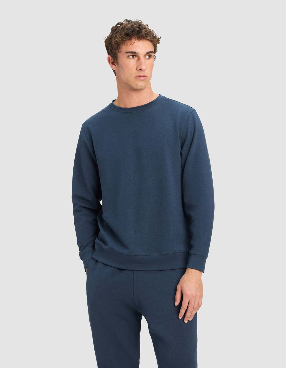 A young man wearing the Cozy Earth Men's StretchTech Crewneck in navy blue stands against a plain light gray background, looking to his left with one hand in his pocket.