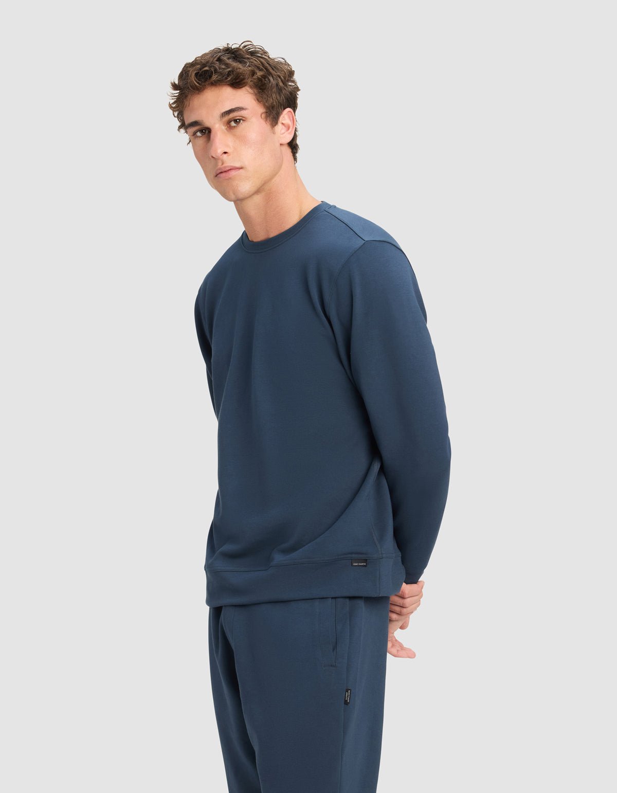 A young man with short, curly brown hair wears a Cozy Earth Men's StretchTech Crewneck in dark blue. He stands against a plain light grey background, looking slightly to the side with his hands behind his back.