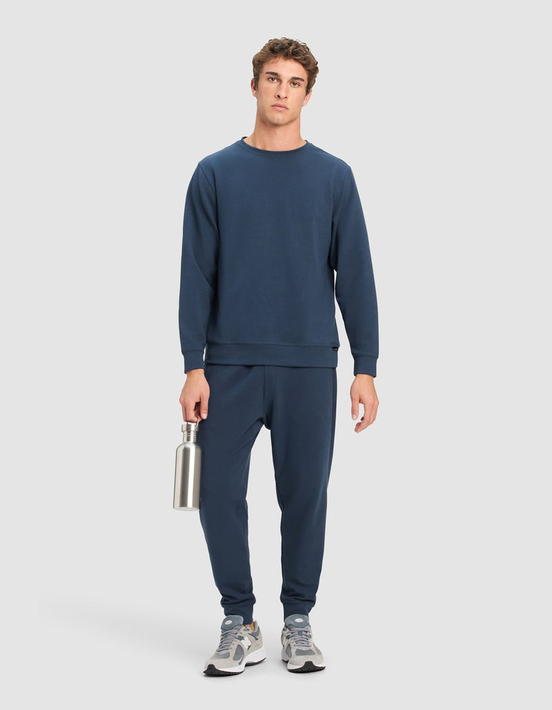 A man faces forward against a light grey background, wearing the Cozy Earth Men's StretchTech Crewneck and matching joggers in navy blue. He holds a silver reusable water bottle and sports grey and white sneakers.