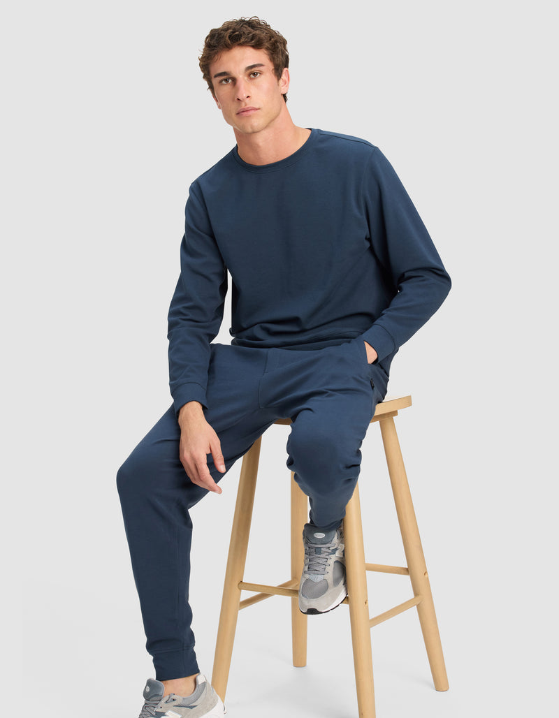 A young man wears the Cozy Earth Men's StretchTech Crewneck & Jogger Set in navy blue, paired with gray sneakers, while seated on a wooden stool against a plain light gray background, looking at the camera with one hand in his pocket.