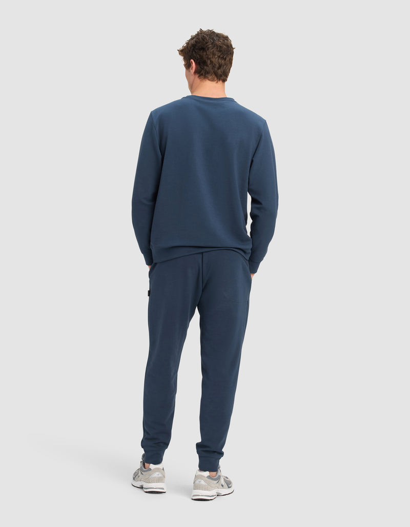 A man faces away, wearing Cozy Earth's Men's StretchTech Crewneck & Jogger Set in navy blue with white sneakers, set against a plain light gray background.