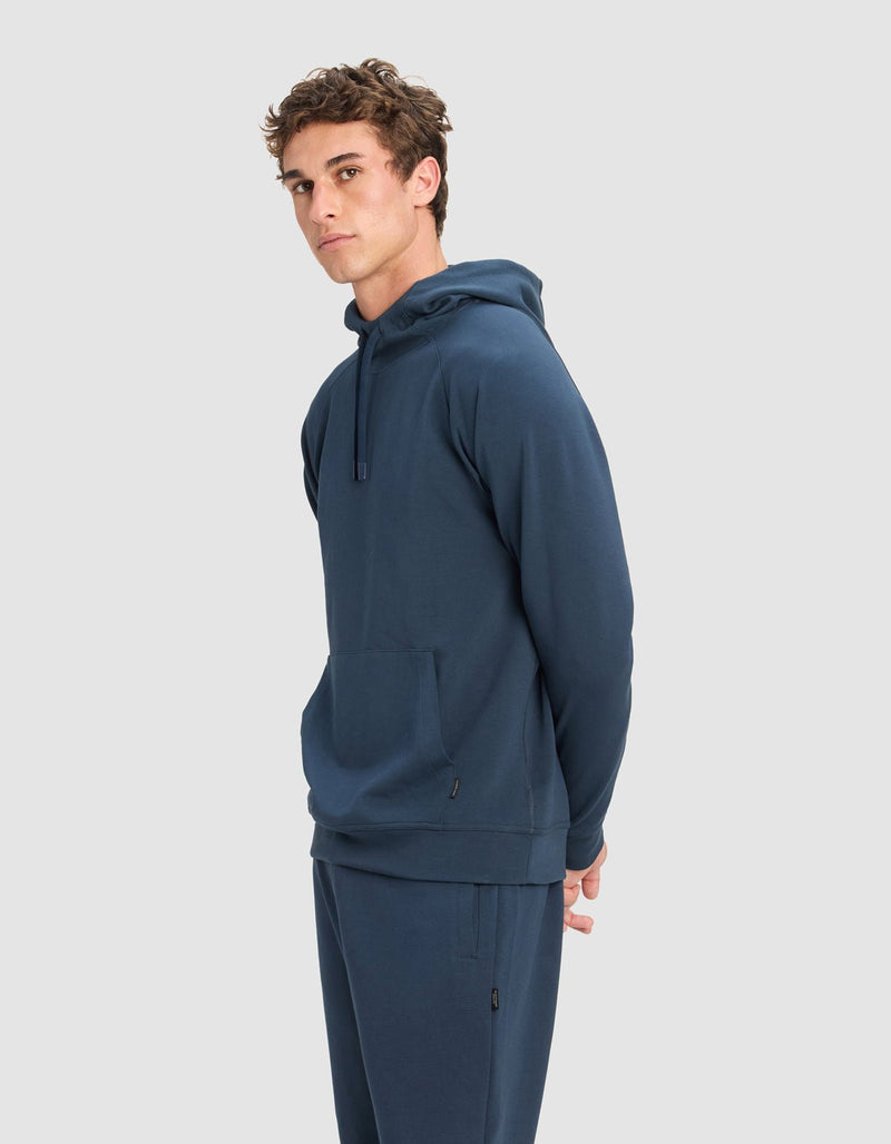 A young man with short, curly brown hair models the Cozy Earth Men's StretchTech Hoodie in blue, paired with matching pants, standing sideways with his hands behind his back against a light gray background.