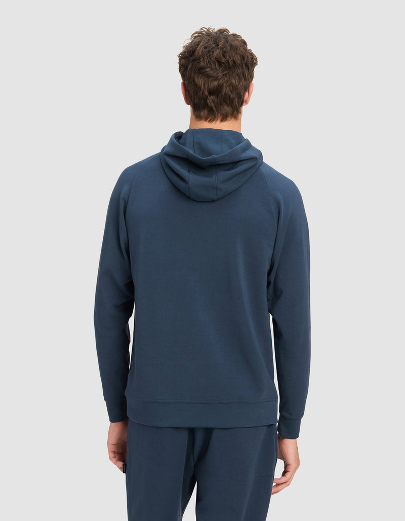 A person with curly brown hair is seen from the back, wearing the Cozy Earth Men's StretchTech Hoodie in dark blue, paired with matching pants, set against a plain light gray background.