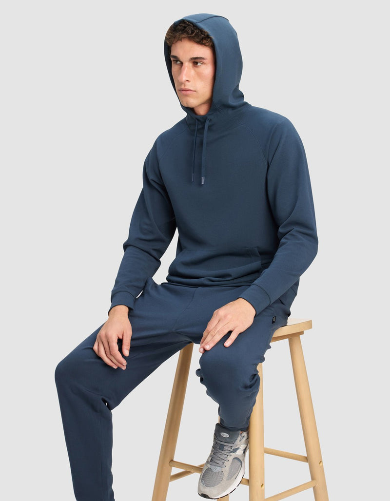 A young man wears the Cozy Earth Men's StretchTech Hoodie, paired with matching sweatpants, sitting on a light wooden stool against a plain light gray background. The hood is up as he looks off to the side with a neutral expression.