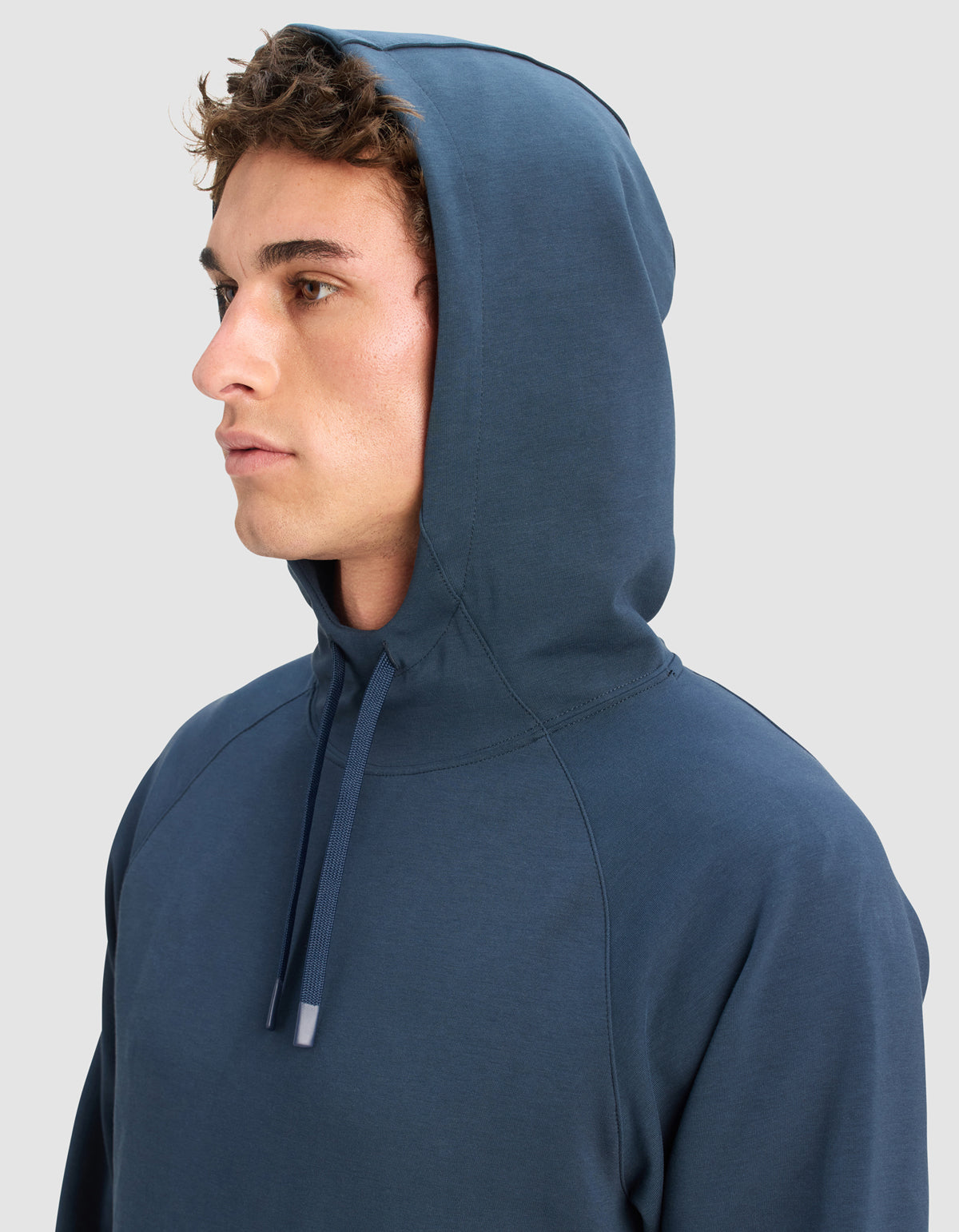 A young man with curly brown hair wears the Cozy Earth Men's StretchTech Hoodie with the hood up, looking to the left against a plain light gray background.