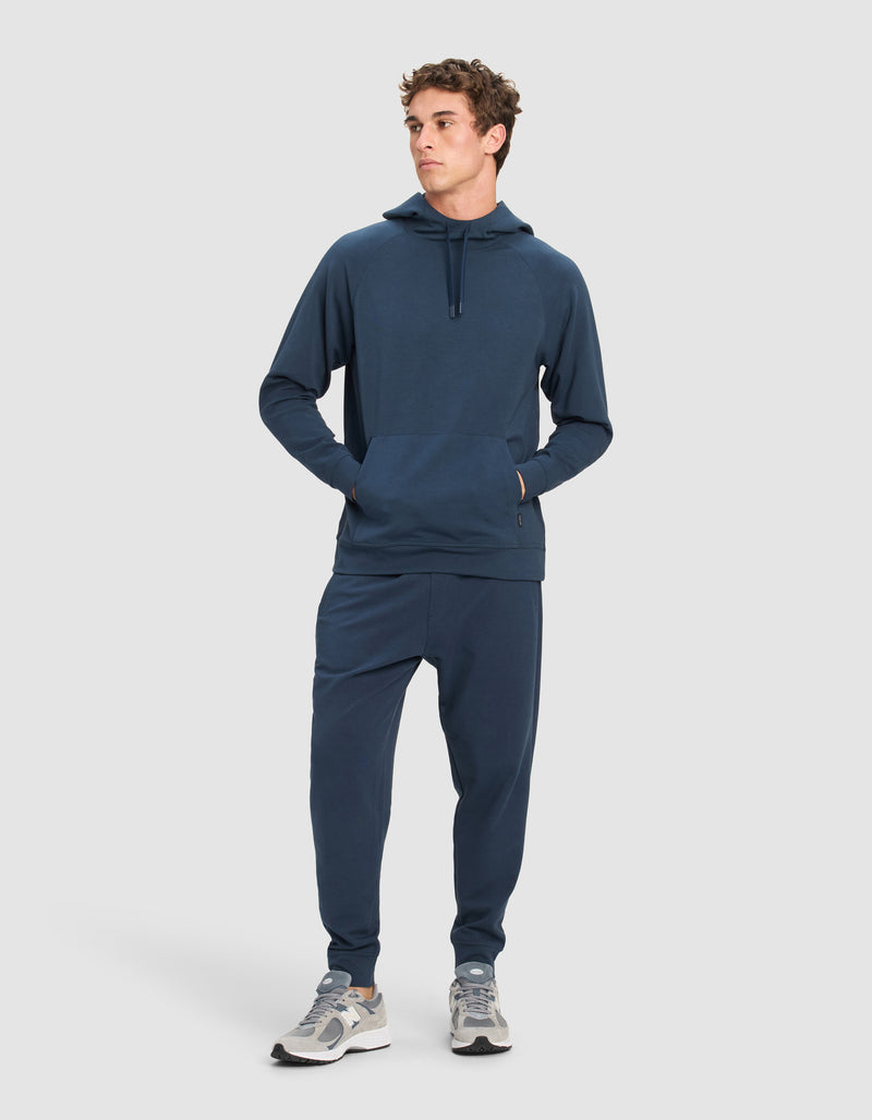 A man stands slightly to the side, wearing the Cozy Earth Men's StretchTech Hoodie & Jogger Set in dark blue with grey and white sneakers, posed against a plain light background.