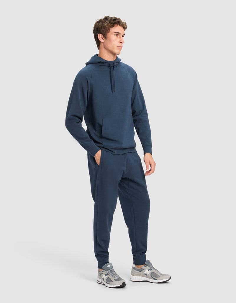 A man wears the Cozy Earth Men's StretchTech Hoodie & Jogger Set in navy, styled with gray and white sneakers, against a plain light gray background.