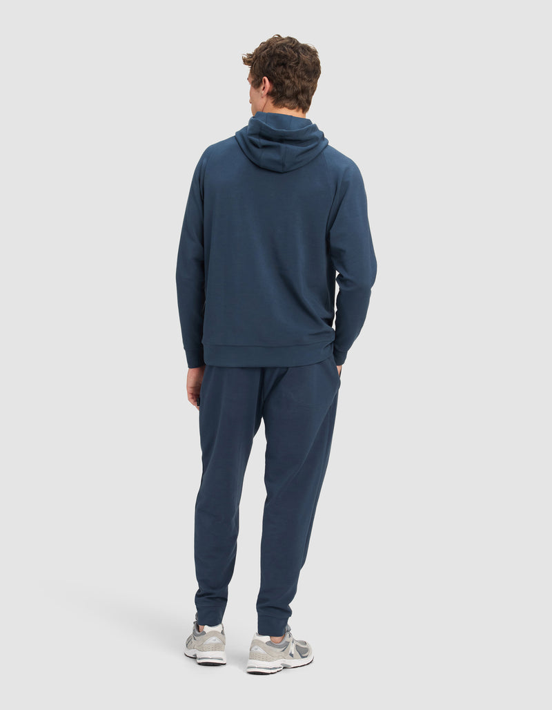 A man faces away, wearing the Cozy Earth Men's StretchTech Hoodie & Jogger Set in dark blue with light gray sneakers, against a plain light background.