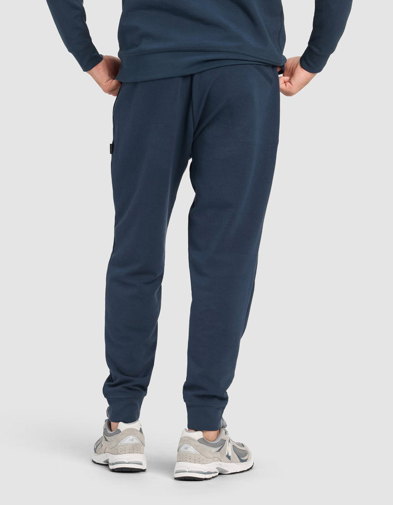 A person shown from the back wearing the Cozy Earth Men's StretchTech Quarter Zip & Jogger Set in navy blue with light gray athletic shoes, standing against a plain light background.