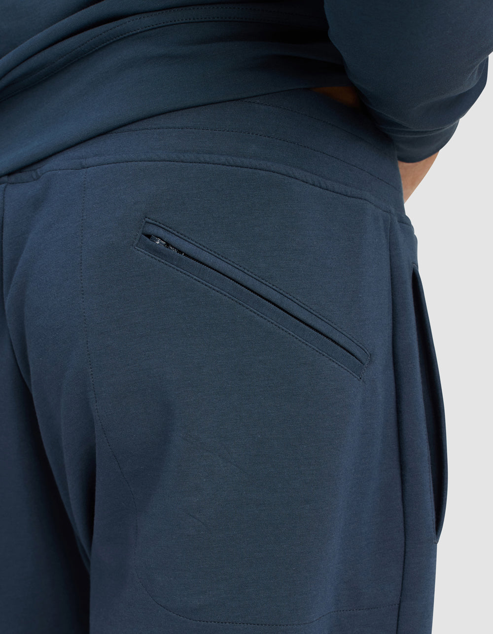 Close-up of a person wearing Cozy Earth Men's StretchTech Quarter Zip & Jogger Set in dark blue, showing a zippered back pocket and one hand partly in the waistband; plain light gray background.