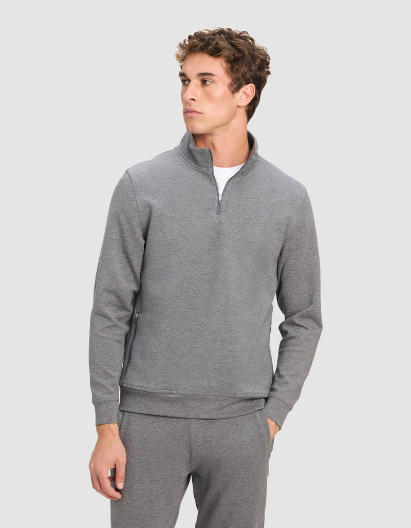 A young man wearing Cozy Earth's Men's StretchTech Quarter Zip and matching pants stands with one hand in his pocket, looking off to the side against a plain light gray background.