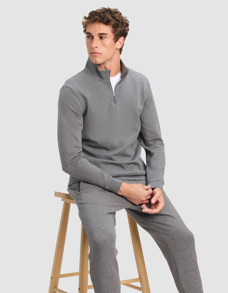 A young man wearing Cozy Earth's Men's StretchTech Quarter Zip in gray with matching pants sits on a wooden stool, looking to the side against a plain light gray background.