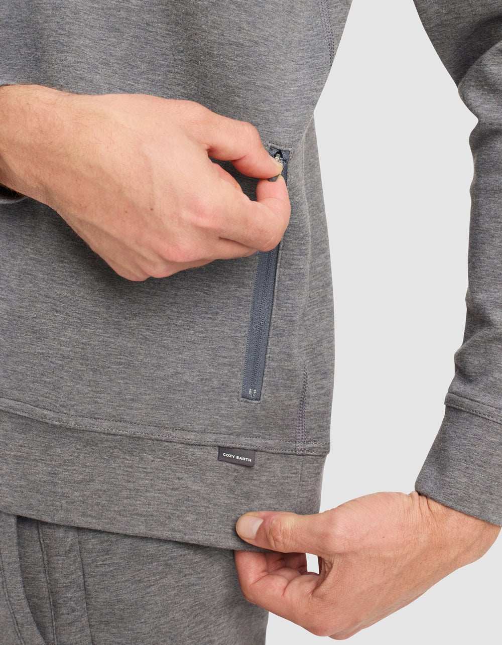 A person in a gray Men's StretchTech Quarter Zip by Cozy Earth is unzipping a vertical side pocket while holding the hem with the other hand. The sweatshirt features a small black "Cozy Earth" label.