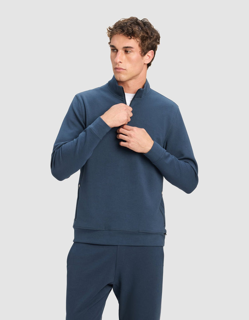A young man stands against a plain light gray background, adjusting the zipper on his Cozy Earth Men's StretchTech Quarter Zip in dark blue, paired with matching pants.