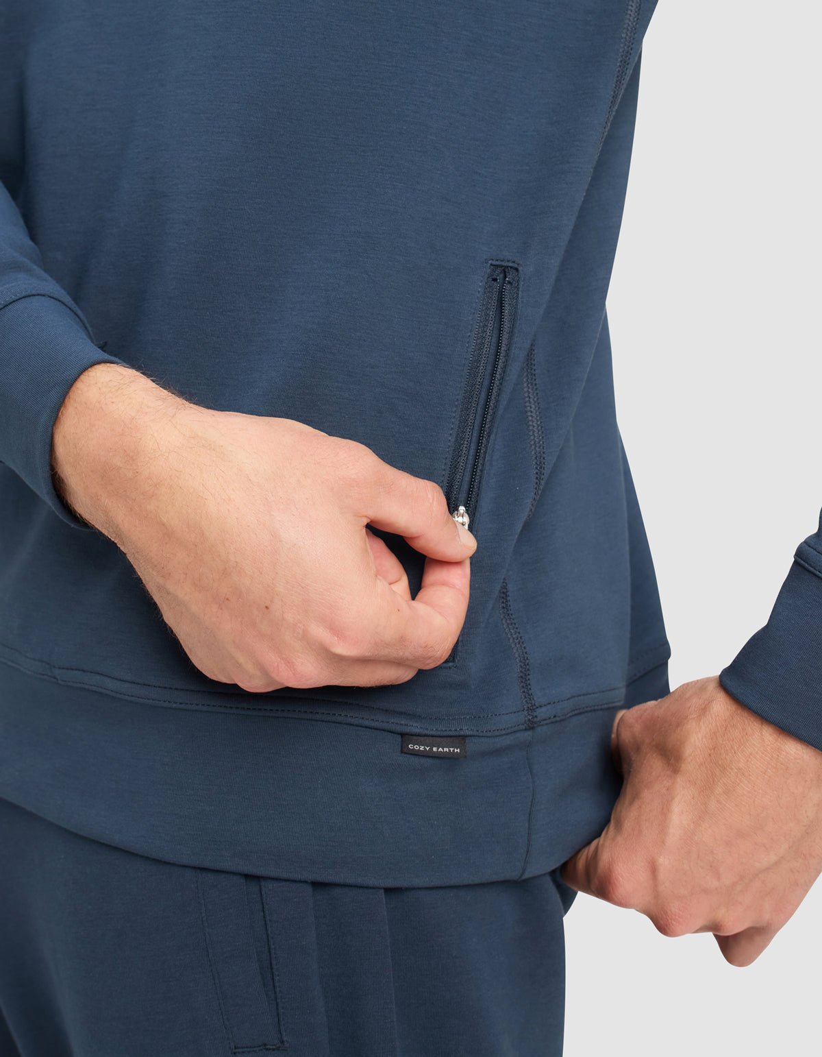 A man wearing Cozy Earth Men's StretchTech Quarter Zip and matching pants is pulling the zipper of a side pocket on the top, set against a plain light gray background.