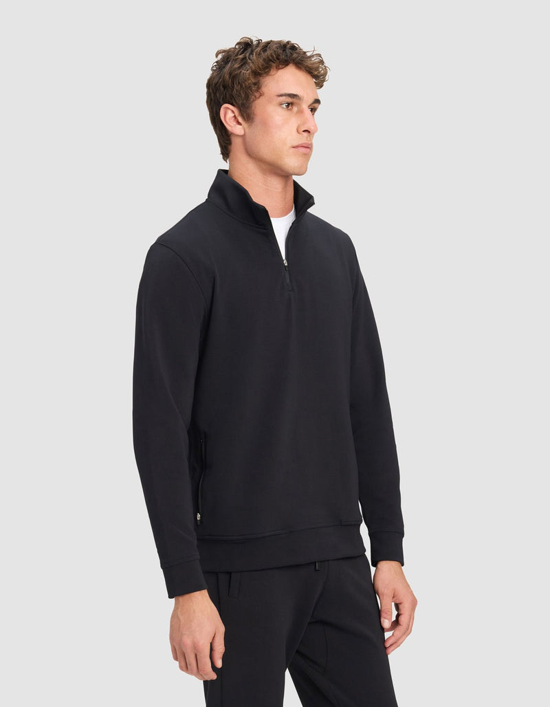 A young man stands slightly turned against a plain light gray background, wearing a Cozy Earth Men's StretchTech Quarter Zip pullover with matching black pants.