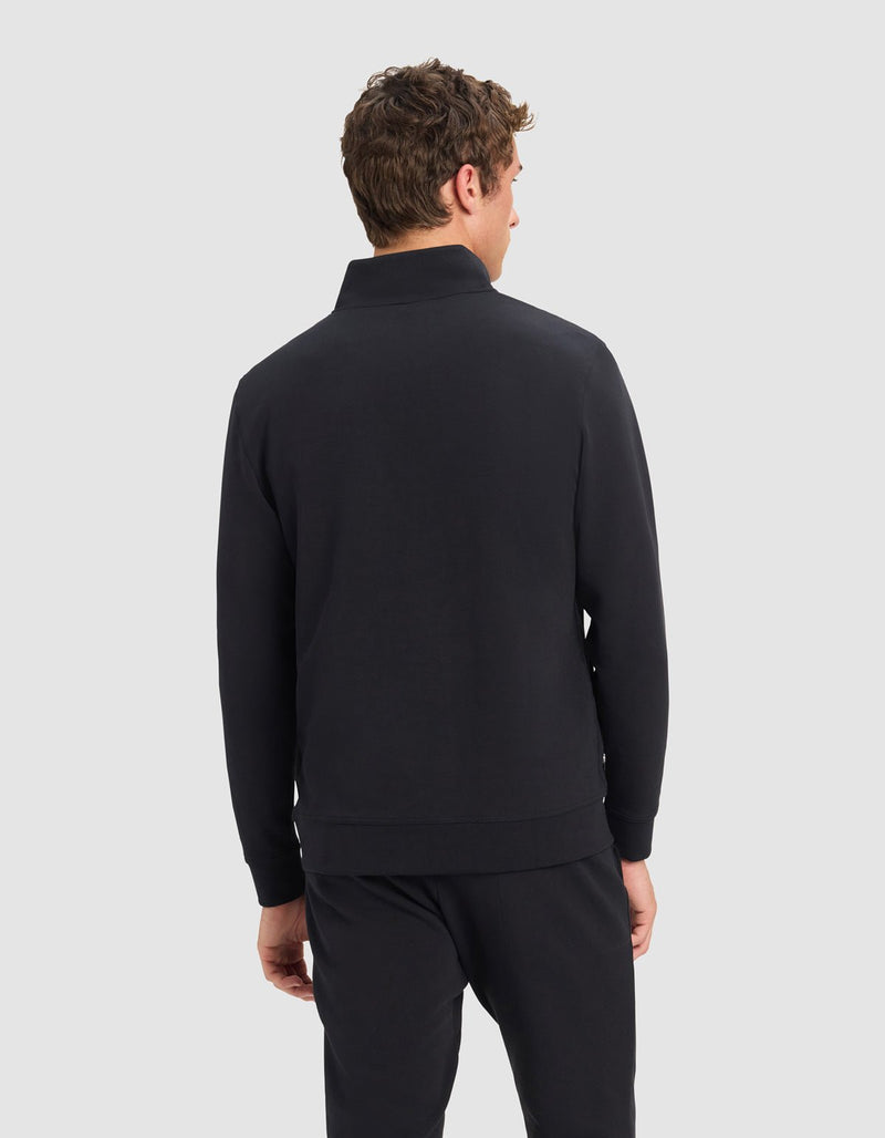 A man with short, curly brown hair is shown from behind, wearing Cozy Earth's Men's StretchTech Quarter Zip in black, paired with matching black pants and standing against a plain light gray background.