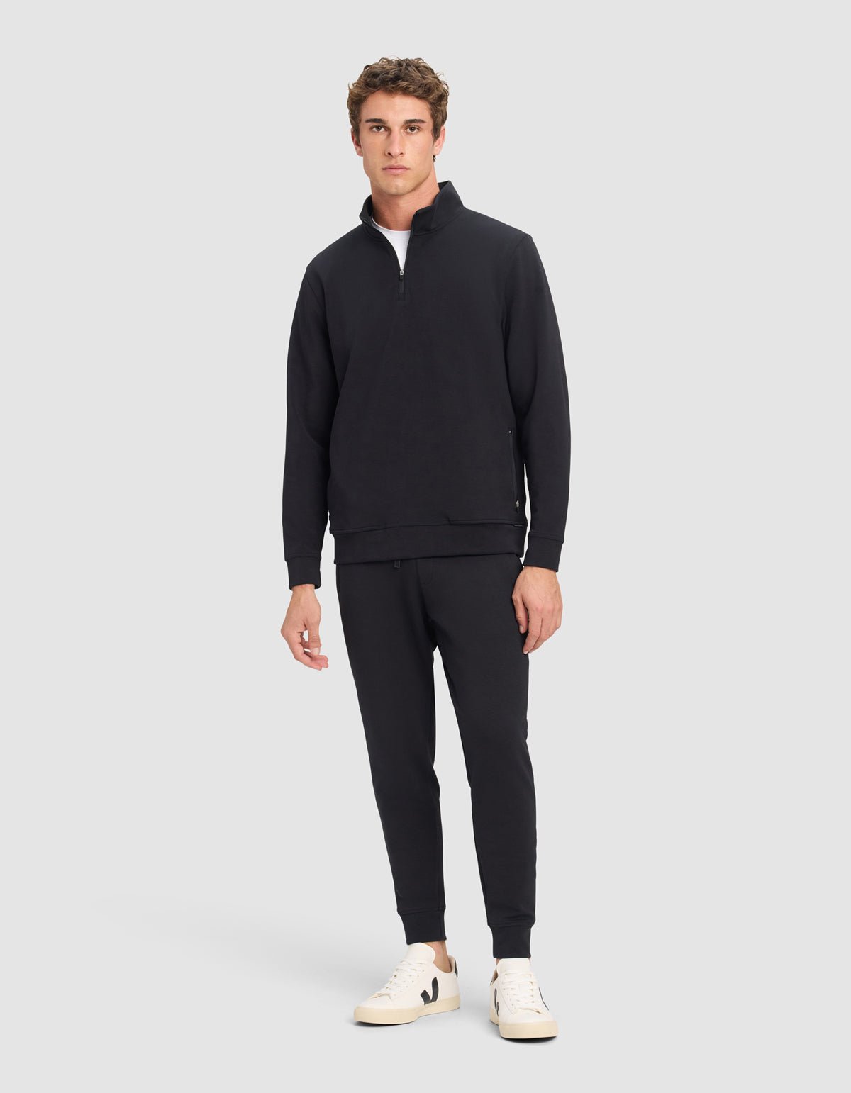 A man stands against a plain background wearing Cozy Earth's Men's StretchTech Quarter Zip in black, matching black joggers, and white sneakers. He faces forward with a neutral expression and relaxed posture.