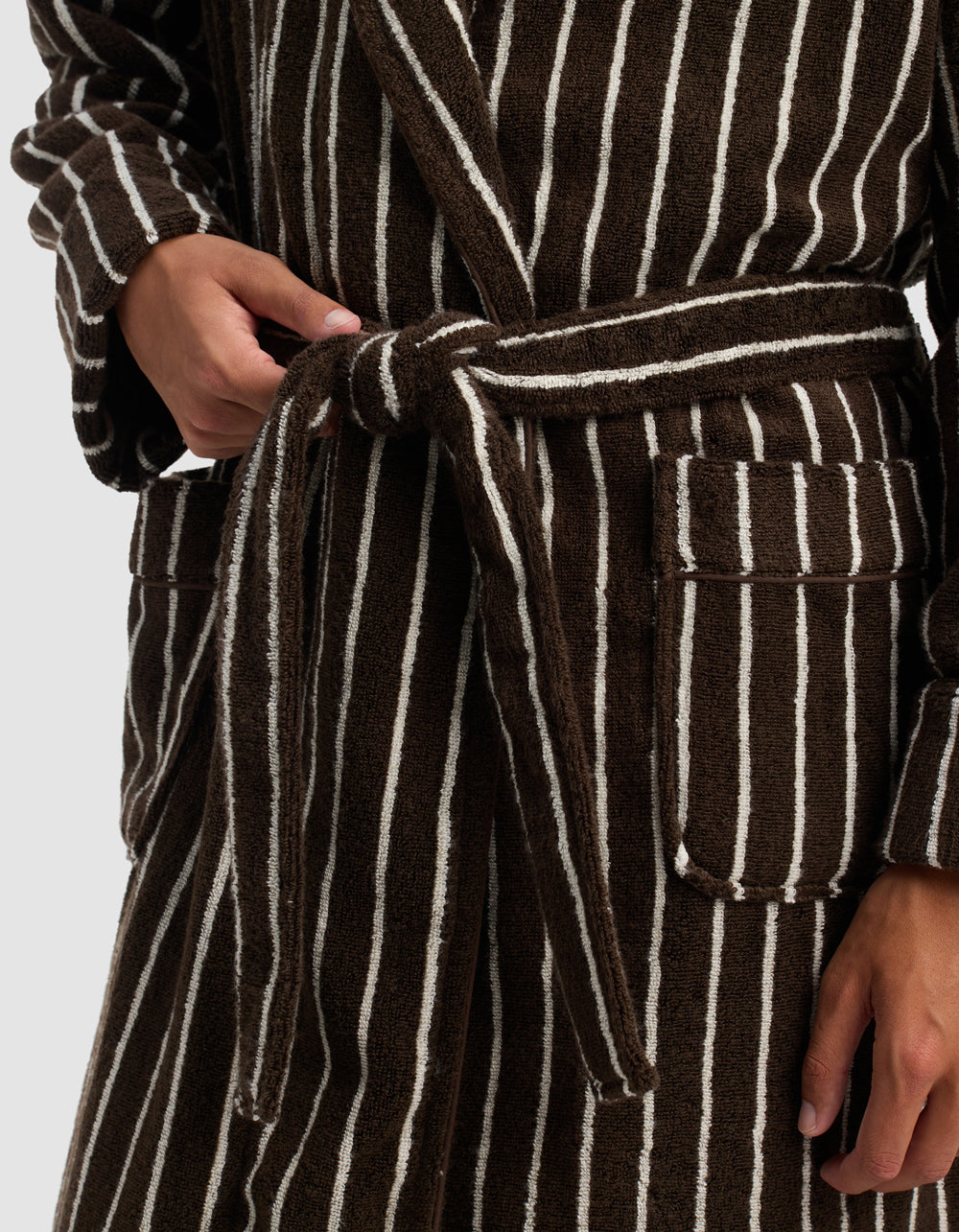 Model wears the Cozy Earth Striped Luxe Bath Robe—brown with white vertical stripes—shown from shoulders to mid-thigh. The robe, tied at the waist with a matching belt, has hands near the belt and pocket.
