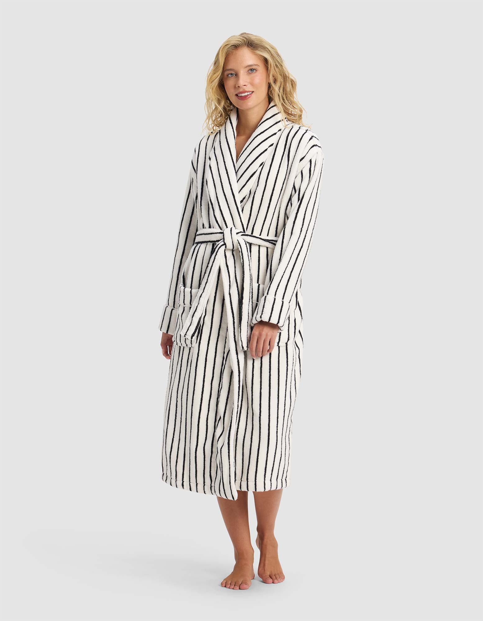 A person stands barefoot in the Striped Luxe Bath Robe by Cozy Earth—an elegant long white robe with vertical black stripes and a tied belt—set against a plain light gray background.