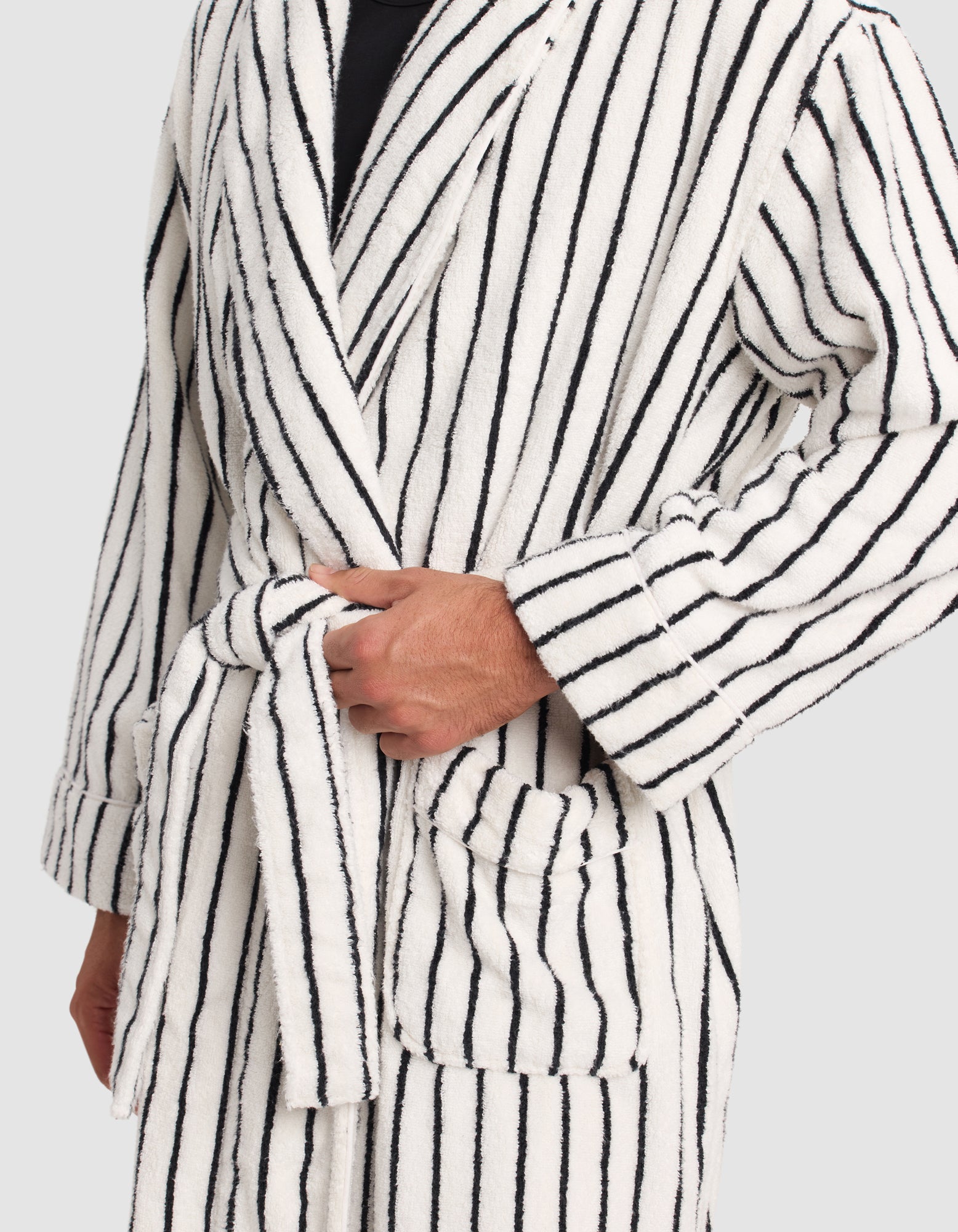 A person wears the Cozy Earth Luxe Bath Robe—white with black vertical stripes, large front pockets, and long sleeves—tying its belt at the waist. Only the torso and one hand are visible.