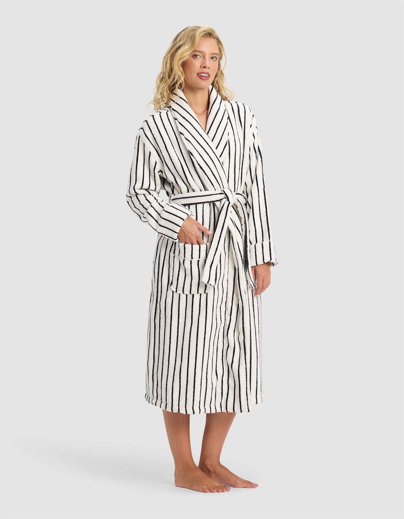 A person stands barefoot in the Cozy Earth Striped Luxe Bath Robe, white with black vertical stripes and a tie waist, hands partially in pockets, against a plain light gray background.